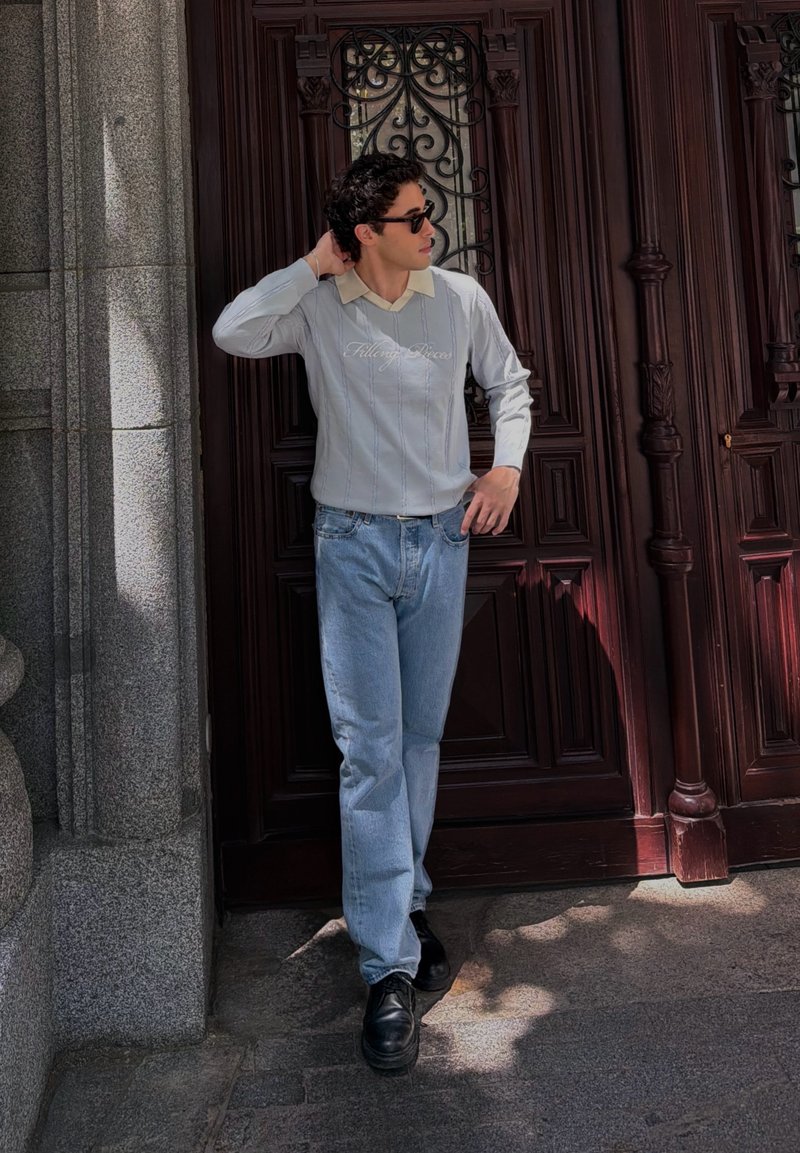 Jeune homme portant des lunettes de soleil, un pull léger et un jean, appuyé contre une grande porte en bois sombre au soleil près d’un mur en pierre.