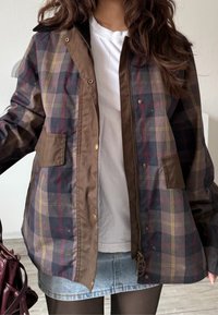 Femme portant une veste à carreaux marron et bleu marine sur une chemise blanche et une jupe en jean, tenant un sac à main rouge foncé avec des cheveux ondulés desserrés.