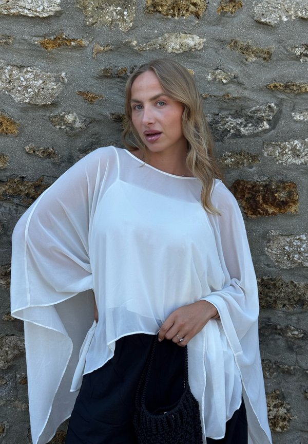 Femme aux cheveux blonds ondulés portant un haut blanc ample et transparent et un pantalon noir, tenant un sac noir tissé, debout contre un mur en pierre.