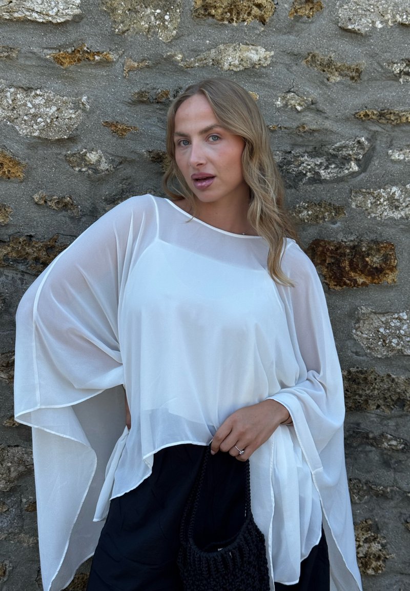 Femme aux cheveux blonds ondulés portant un haut blanc ample et transparent et un pantalon noir, tenant un sac noir tissé, debout contre un mur en pierre.