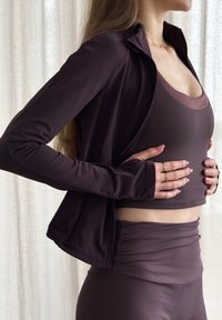 Costum sportiv mov închis, format dintr-o jachetă cu fermoar, un top fără mâneci și colanți cu talie înaltă cu detaliu ruching, confecționat dintr-un material neted și elastic.