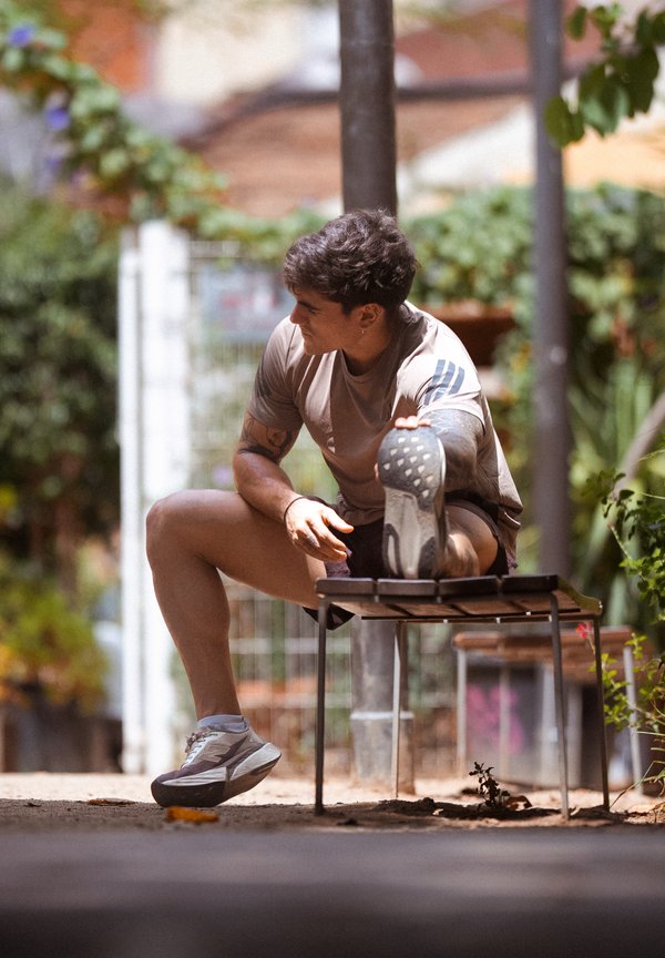 Homme en tenue de sport étirant une jambe sur un banc en plein air avec de la verdure et une clôture en arrière-plan.
