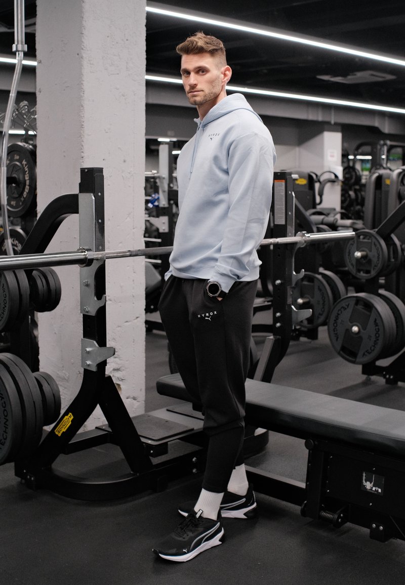 Jeune homme en tenue de sport debout près d'une barre chargée de poids sur un support à squats dans une salle de sport moderne.