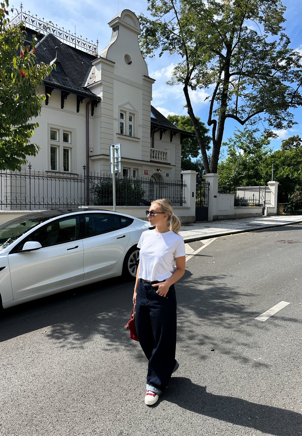 Bela kratica majica, temni široki hlače in bele superge s rdečimi poudarki. Drži majhno rdečo torbico. Urbani okolje z hišo in drevesom.