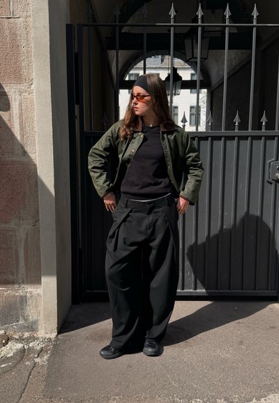 Chaqueta de algodón verde, suéter negro y pantalones oversized negros. Gafas de sol oscuras y zapatillas negras completan el look. Texturas mate, ajuste relajado.