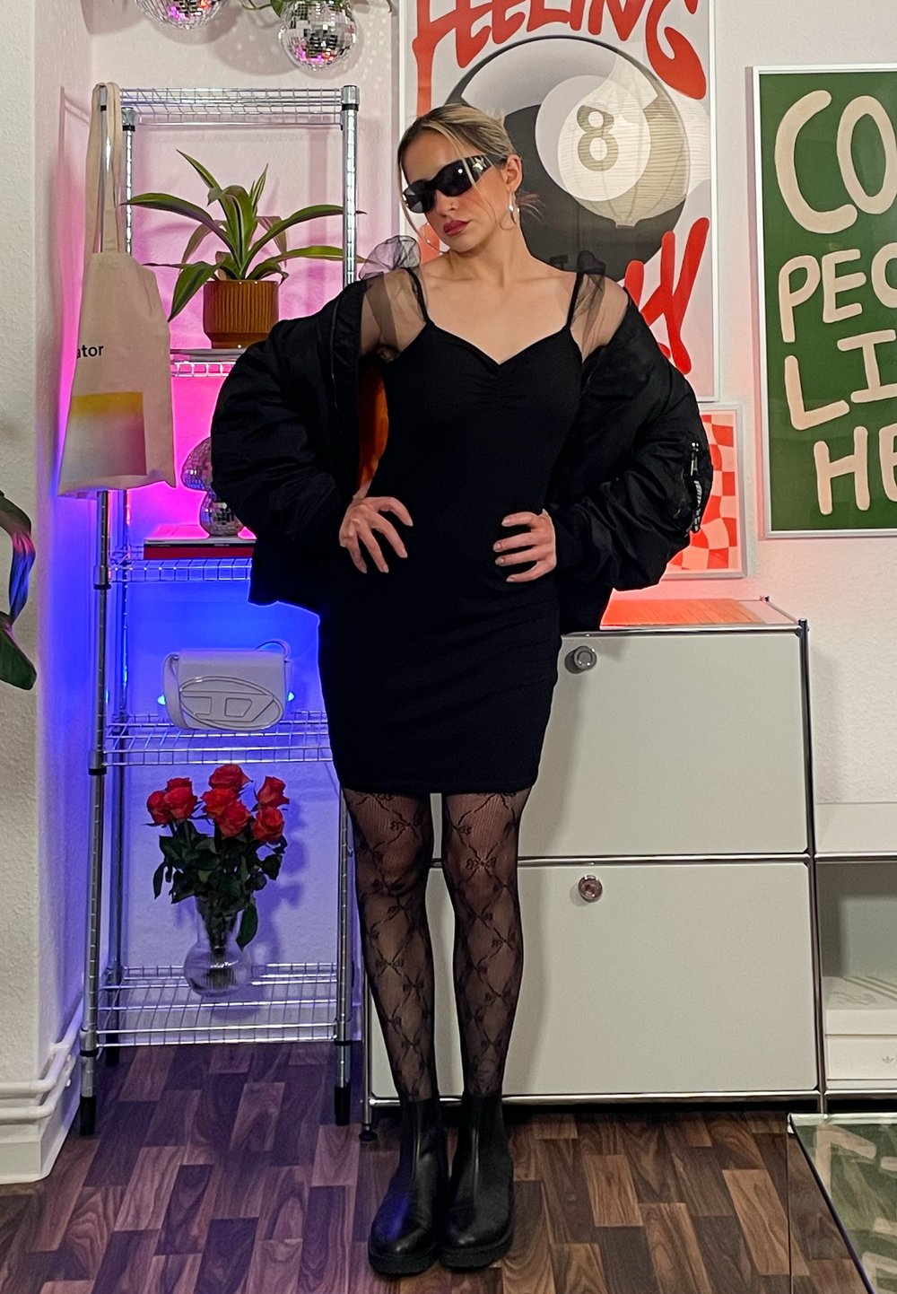 Black fitted dress with sheer accents, oversized black bomber jacket, patterned tights, and black ankle boots. Accessories include sunglasses and hoop earrings.