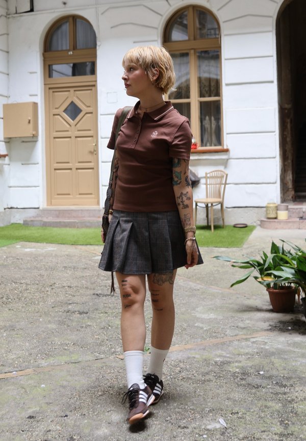 Persona joven con el pelo corto y rubio caminando por un patio, lleva un polo marrón, falda de cuadros, calcetines blancos y zapatillas negras.
