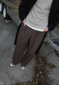 Pantaloni ampi a righe marroni abbinati a un maglione grigio texturizzato indossato sopra una camicia bianca, completati da sneakers distintive con un dettaglio nero.