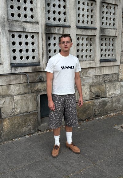 Camiseta blanca con el texto "SUNNEI", pantalones cortos grises con estampado, calcetines blancos y zapatos marrones con cordones, todo esto frente a una pared de piedra texturizada.
