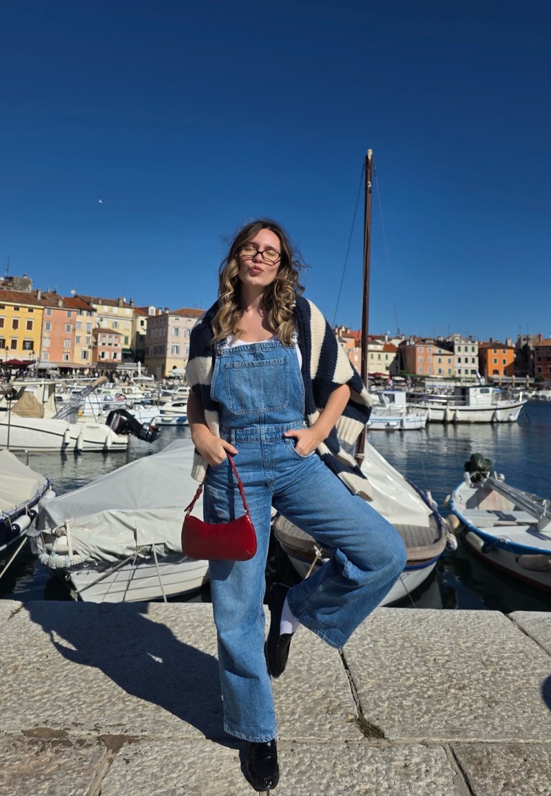 Blue denim overalls with wide legs, paired with a black and white striped sweater and a red handbag. Background includes boats and colorful buildings.