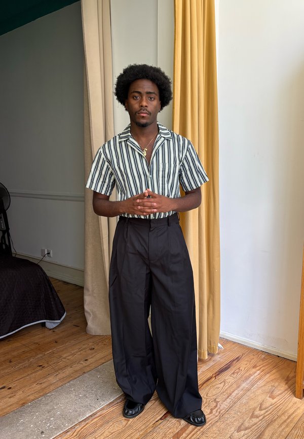 Young man with short curly hair wearing a striped short-sleeve shirt and wide black trousers stands indoors with hands clasped.