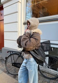 Personne en veste marron et capuche beige ajustant la capuche près d'un vélo noir contre un bâtiment avec une grande fenêtre.