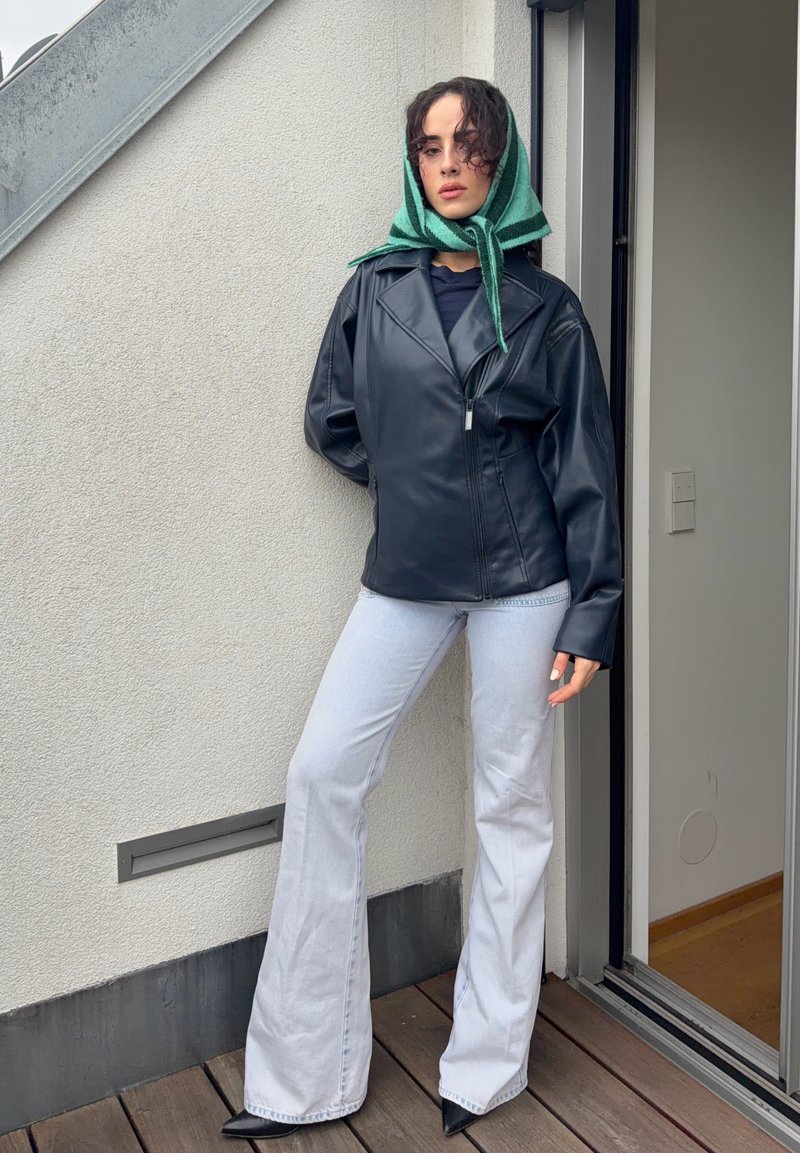Person wearing a black leather jacket, white flared jeans, black pointed shoes, and a green striped headscarf, standing on a wooden deck near a door.