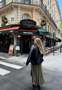 Kahvilan sisäänkäynti, jossa on retro-tyylinen kyltti, vihreä ja punainen markiisi sekä ulkona istumapaikkoja. Mustaan nahkatakkiin ja oliivinvihreään hameeseen pukeutunut henkilö kävelee ohi.