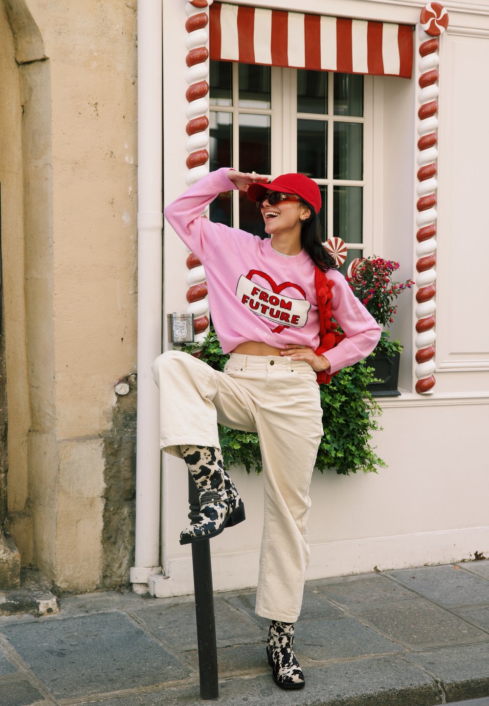 Pulover roz cu textul "DIN VIITOR", pantaloni cremoși cu picioare largi, cizme cu un model alb-negru. Fundalul prezintă o fereastră și accente decorative.