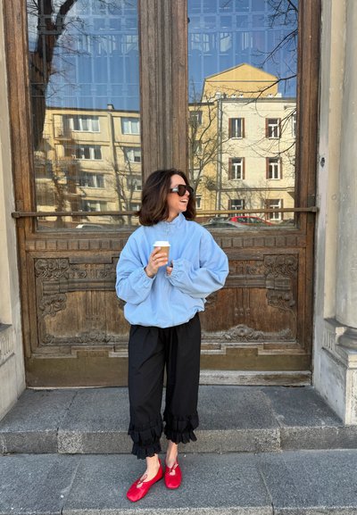 Mujer con gafas de sol, chaqueta azul, pantalones negros y zapatos rojos, sosteniendo una taza de café de pie en unos escalones de piedra frente a una gran puerta de madera.