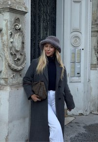 Hall triibuline mantel, tekstuuriga hall müts, tumesinine kõrge kaelusega pluus ja valged püksid. Käes pruunikasmustriline käekott. Ornamentidega kivitaust.