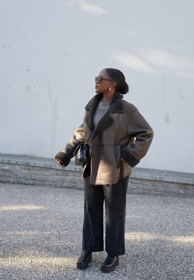 Chaqueta de piel con forro de borreguito marrón y puños negros, combinada con pantalones de pierna ancha oscuros y botas de plataforma negras. Accesorio con un bolso pequeño.