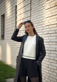 Čierny kožený dlhý kabát cez biele tričko s čiernym pruhom, skombinovaný s čiernymi šortkami. Modelka drží slnečné okuliare a pózuje proti tehlovej stene.