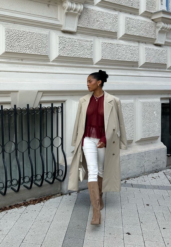 Cappotto beige doppiopetto, top trasparente bordeaux con pieghe, pantaloni bianchi e stivali al ginocchio color cammello. Rete metallica nera sullo sfondo.