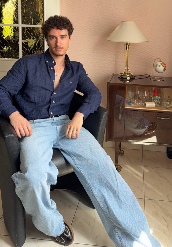 Junger Mann mit lockigem Haar, der ein dunkelblaues Hemd und hellblaue Jeans trägt, sitzt in einem schwarzen Sessel neben einem Holzschrank mit einer Lampe.