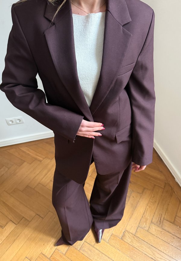 Person trägt einen dunkelbraunen Anzug und einen weißen Pullover, steht auf einem Holzboden und richtet mit einer Hand die Jacke, sichtbare spitze Schuhe.