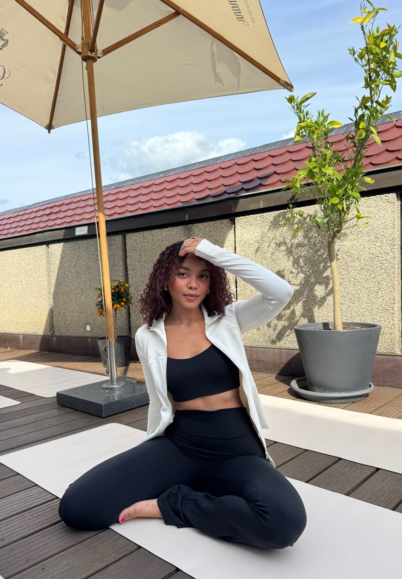 Femme en tenue de sport noire et veste blanche assise jambes croisées sur un tapis de yoga sous un parasol de terrasse sur une terrasse en bois.