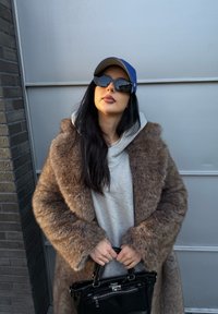 Cappotto in pelliccia sintetica marrone, felpa grigia con cappuccio, occhiali da sole neri, berretto azzurro e una borsa nera lucida. Presenta maniche lunghe e una vestibilità rilassata.