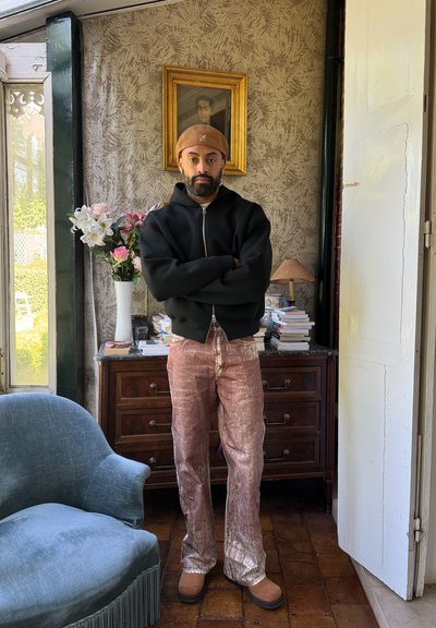 Sudadera negra con cremallera, pantalones anchos brillantes rosa, botas marrones. Jarrón floral al fondo, papel tapiz estampado, muebles de madera.