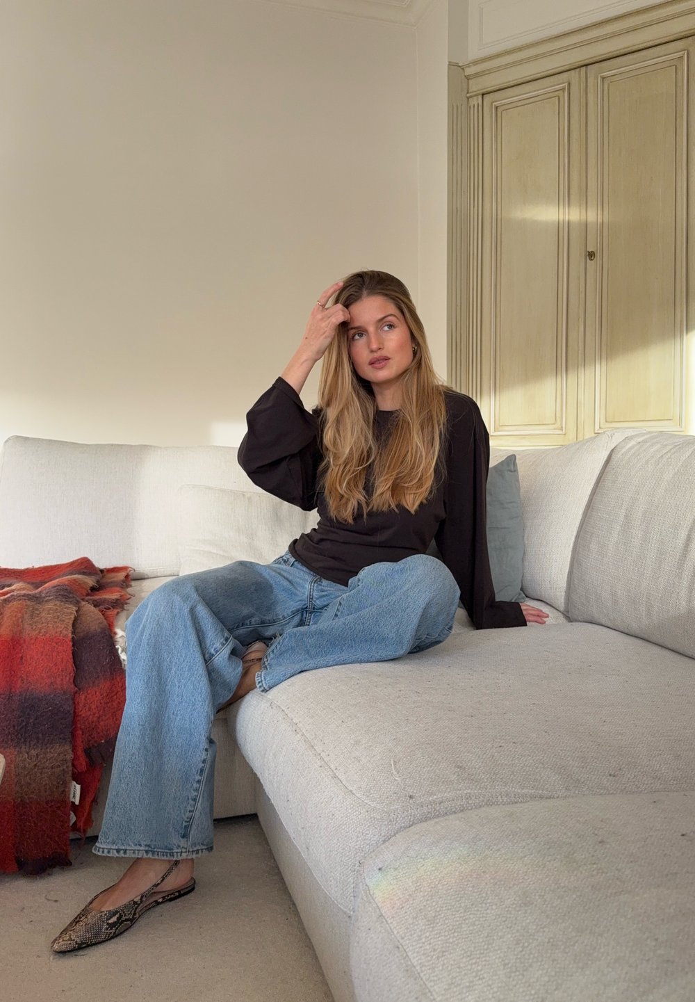 Haut noir à manches longues, jean bleu ample, et ballerines à motif serpent portés en position assise sur un canapé en tissu gris clair avec une couverture à carreaux rouge et marron.