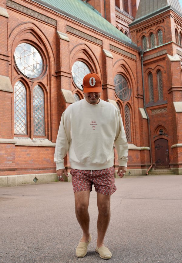 Homme portant un sweat-shirt blanc, un short à motifs, des chaussures beige sans lacets et une casquette orange marchant à l'extérieur près d'un bâtiment en brique rouge avec des fenêtres en arc.