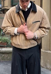 Žametna jakna v bež barvi s črnim usnjenim ovratnikom in žepom na prsih z zadrgo, oblečena čez črn pulover in kombinirana z hlačami z ozkimi črtami.