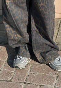Black denim trousers adorned with a brown leopard print, showcasing a wide-leg design. Grey sports shoes with textured soles are also visible.