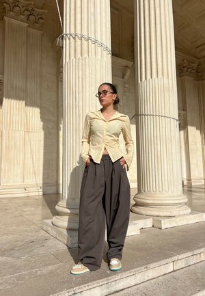 Jasnożółta koszula w paski z kołnierzykiem i długim rękawem, w połączeniu z szerokimi brązowymi spodniami i butami z niebieskimi akcentami. Klasyczne kolumny architektoniczne w tle.