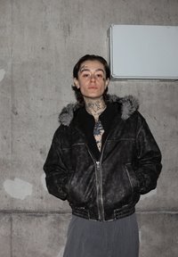 Črna bomber jakna iz umetnega usnja z na krzno obloženim kapuco, zadrgo spredaj in stranskimi žepi. Oblečena prek črne majice in hlač z navpičnimi črtami.