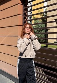 Lys beige, kort jakke med store knapper, brukt over en svart topp og svarte joggebukser med hvite sidestriper. Solfylt bylandskap.