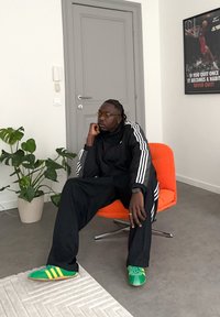 Ensemble de survêtement noir avec des rayures blanches, lunettes jaunes, baskets vertes et jaunes, assis sur une chaise orange, en intérieur avec une plante.