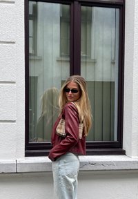 Burgundská kožená zkrácená bunda s kostkovaným detailem na kapse, nošená přes světle modré džíny. Viditelné odrazy ve okně vzadu.