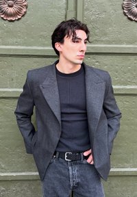 Blazer de lana gris con hombros estructurados, combinado con una camiseta negra ajustada y vaqueros oscuros. Presenta una solapa clásica y una tela texturizada.