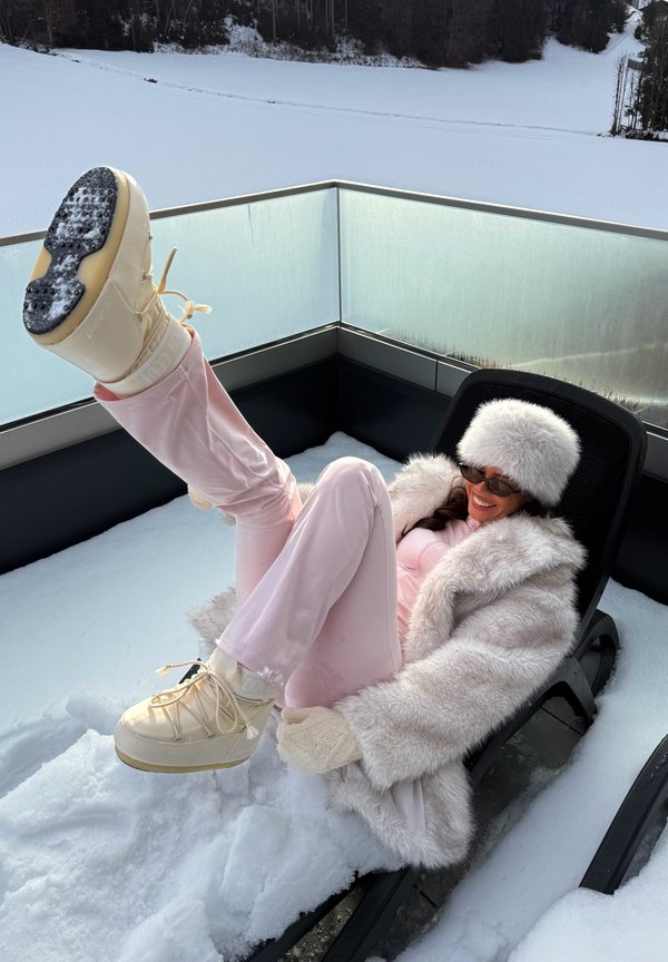 Pantalones de terciopelo rosa claro, abrigo y sombrero blancos y mullidos, botas beige con cordones, sentado en una silla de descanso negra con nieve de fondo.