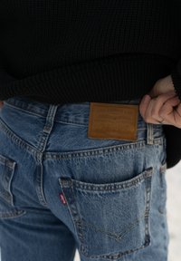 Personne portant un jean Levi's bleu et un pull noir, vue de dos, ajustant la taille du pantalon avec les mains.