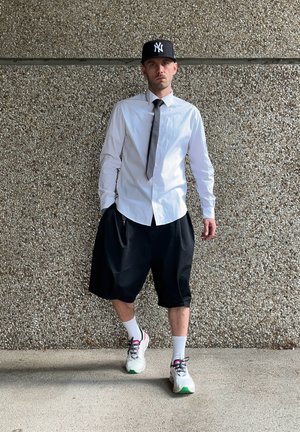Camisa blanca de botones, pantalones cortos oscuros de gran tamaño, corbata gris, gorra negra con logo, calcetas blancas y zapatillas con acentos coloridos. Fondo texturizado.