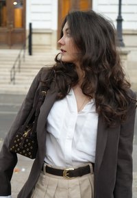 Smeđi sako, bijela košulja, bež plisirane hlače i tamna torbica sa zlatnim detaljima. Valovita kosa i minimalan nakit zaokružuju izgled.