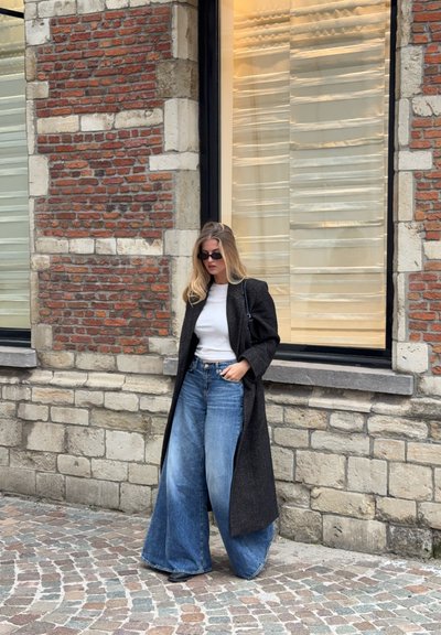 Abrigo largo negro sobre una blusa blanca, combinado con unos vaqueros de pierna ancha azules. Textura suave, diseño clásico, con un fondo de pared de ladrillo.