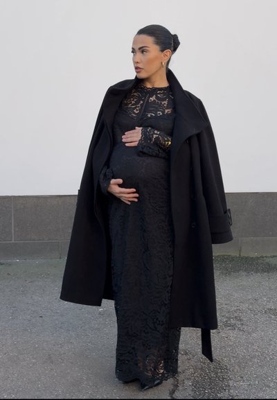 Mujer embarazada con un vestido de encaje negro y abrigo, de pie al aire libre, sosteniendo suavemente su vientre y mirando hacia un lado.