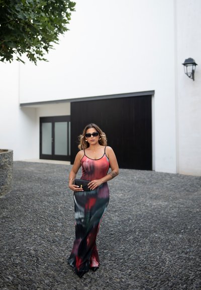 Vestido maxi con tirantes finos que presenta un patrón en degradé en rojo, negro y verde. Textura suave, acompañado de un bolso de mano negro.