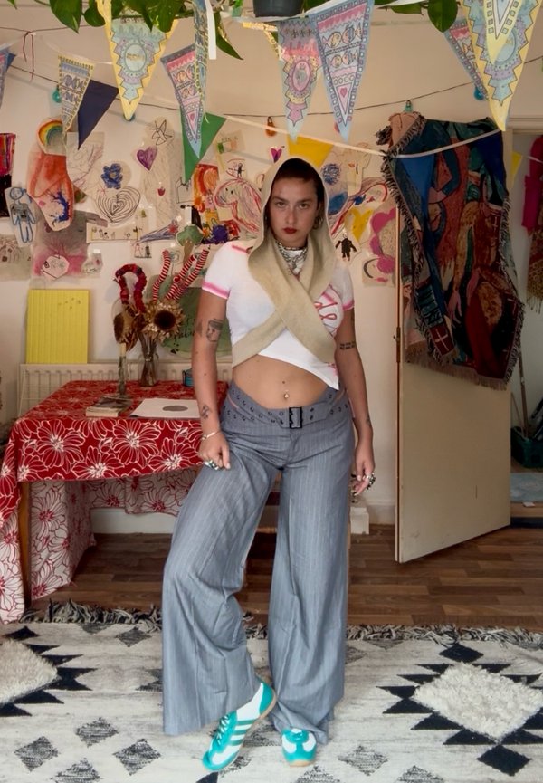 Une personne pose dans une pièce décorée, vêtue d’un crop top blanc, d’un pantalon gris ample, de baskets turquoise et d’une écharpe beige croisée sur le torse.