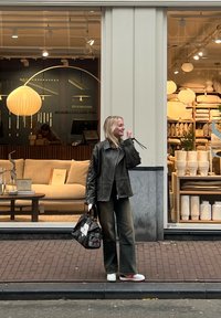 Mörk läderjacka, olivgröna vida byxor, vita sneakers med röda accenter och en svart väska med blommotiv. Butiksinteriör synlig.