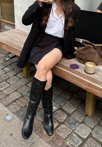 Schwarze Leder kniehohe Stiefel mit spitz zulaufenden Zehen, getragen zu einem schwarzen Rock und einem weißen Hemd, sitzend auf einer Holzbank neben einer braunen Tasche.