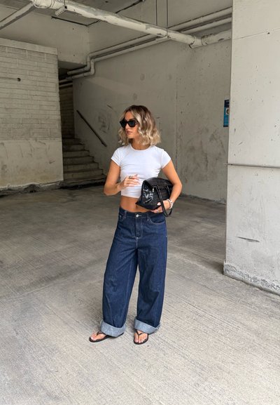Camiseta blanca ajustada, jeans de pierna ancha azul oscuro con puños enrollados, bolso negro y gafas de sol. Fondo minimalista con texturas de concreto.